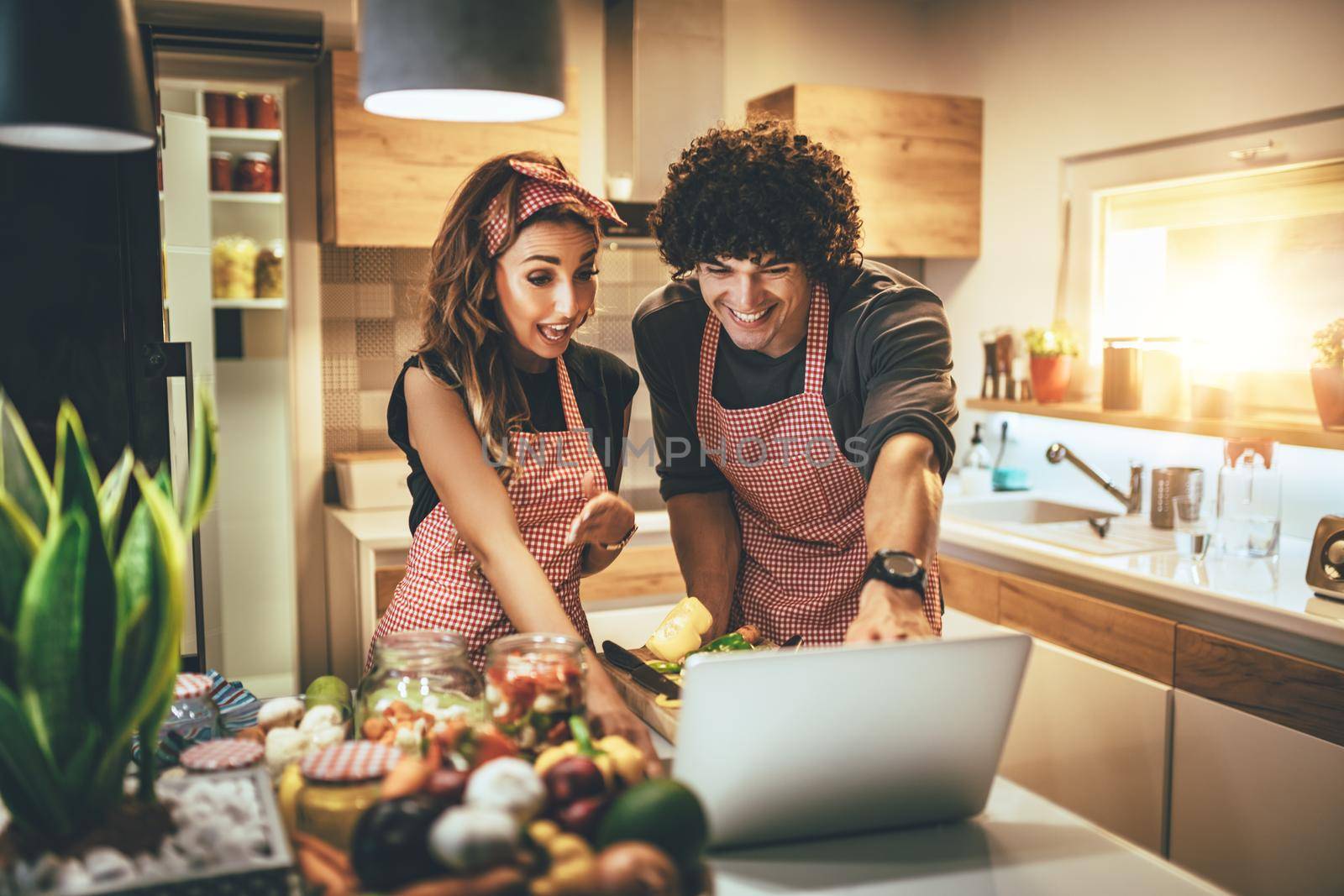 Young happy couple is enjoying and preparing healthy meal in their kitchen and reading recipes on the laptop.