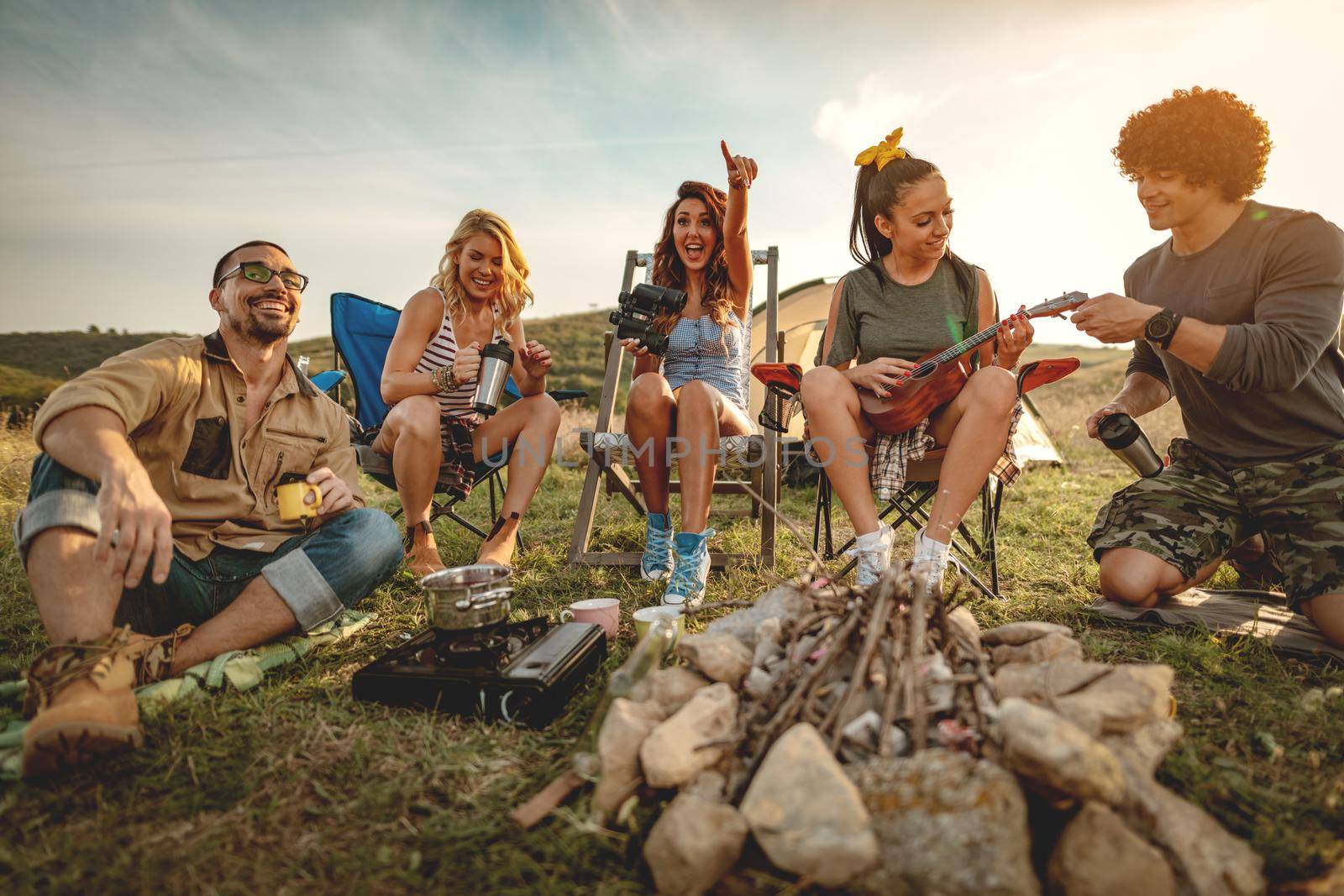 Young people have a good time in camp in nature. They're resting, laughing and singing with music from ukulele, happy to be together.