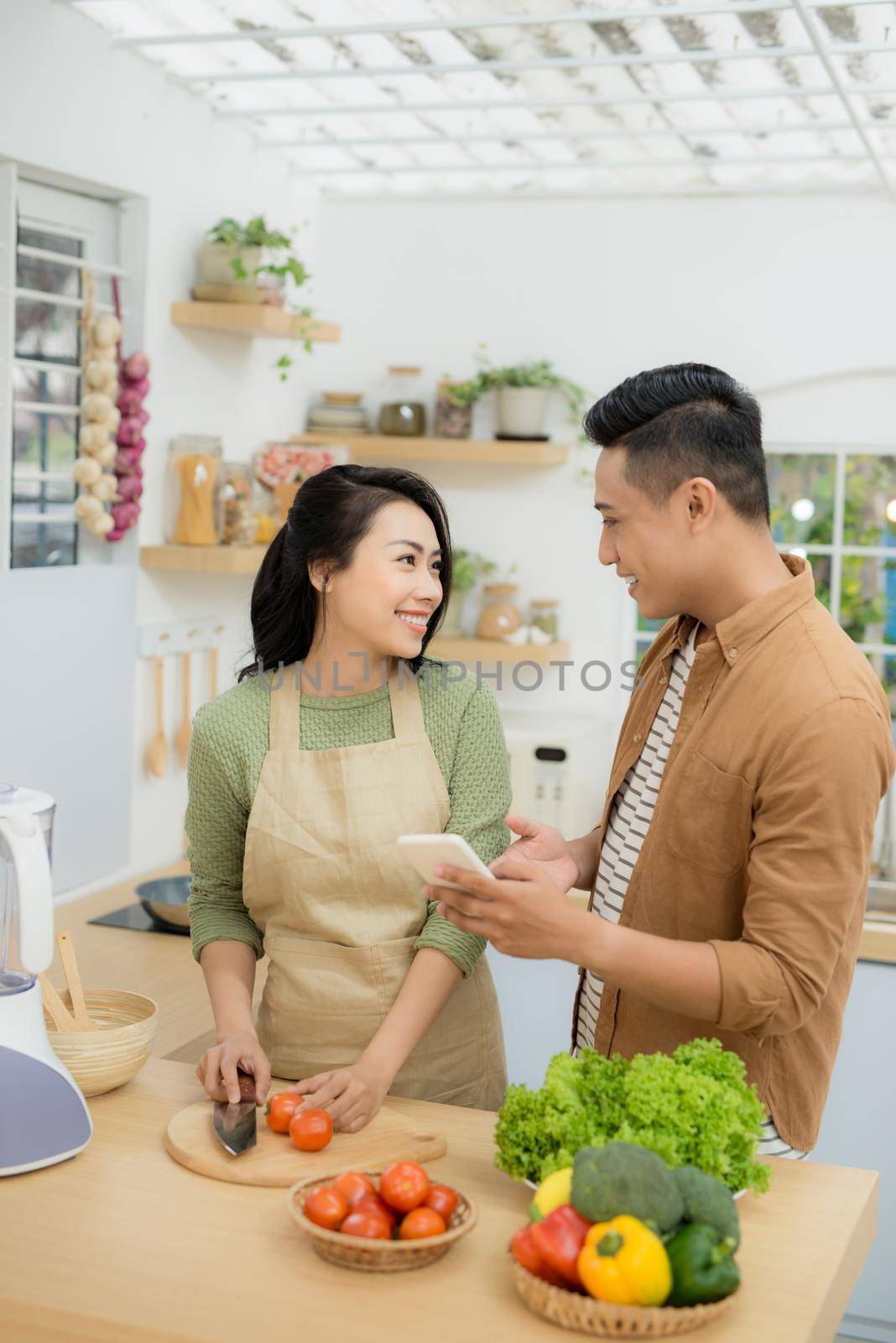 Beautiful young couple is using a cellphone and smiling while cooking in kitchen at home by makidotvn