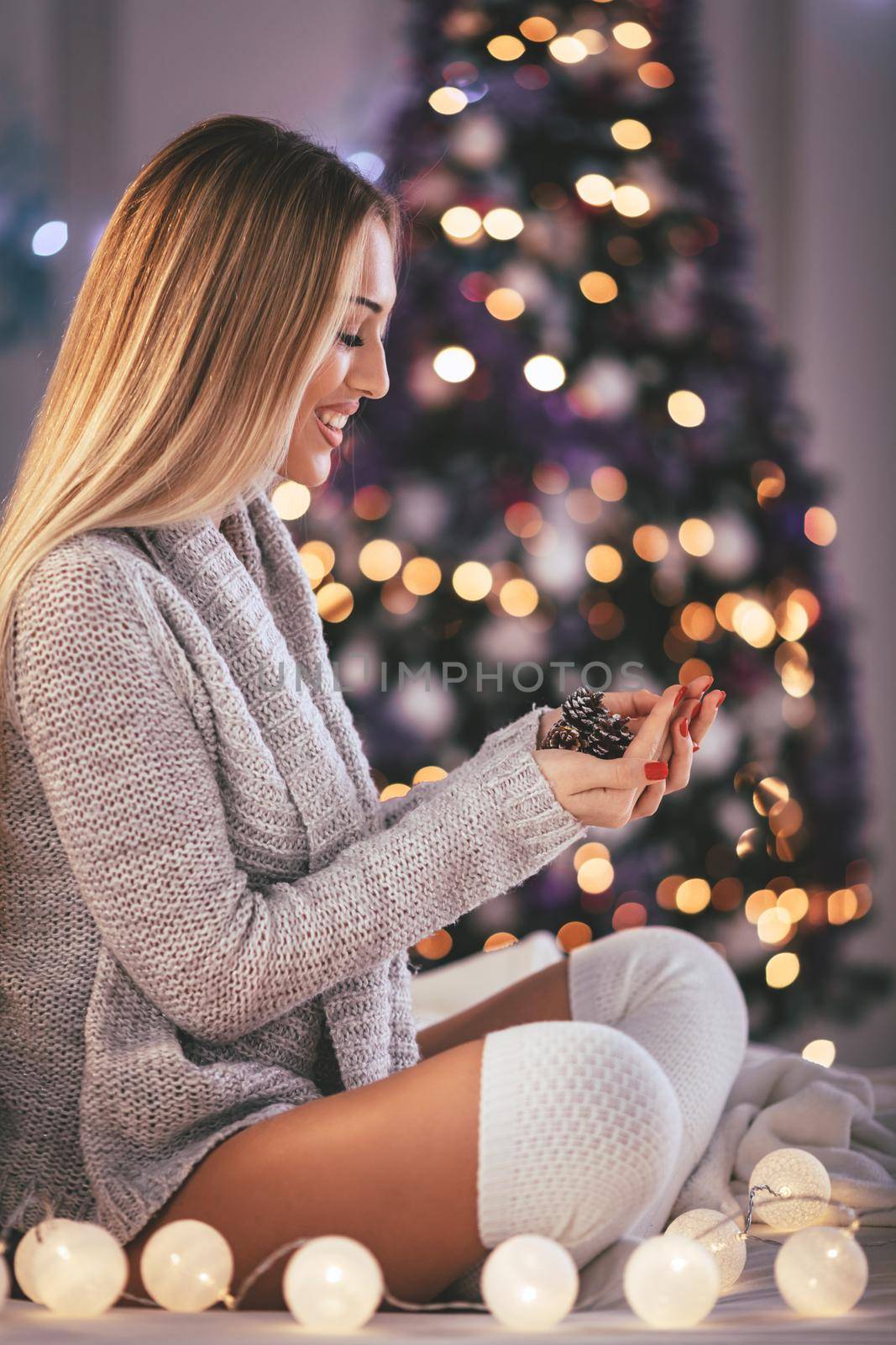 A cute young smiling woman holding pinecones in her hands surrounded with Christmas lights. 