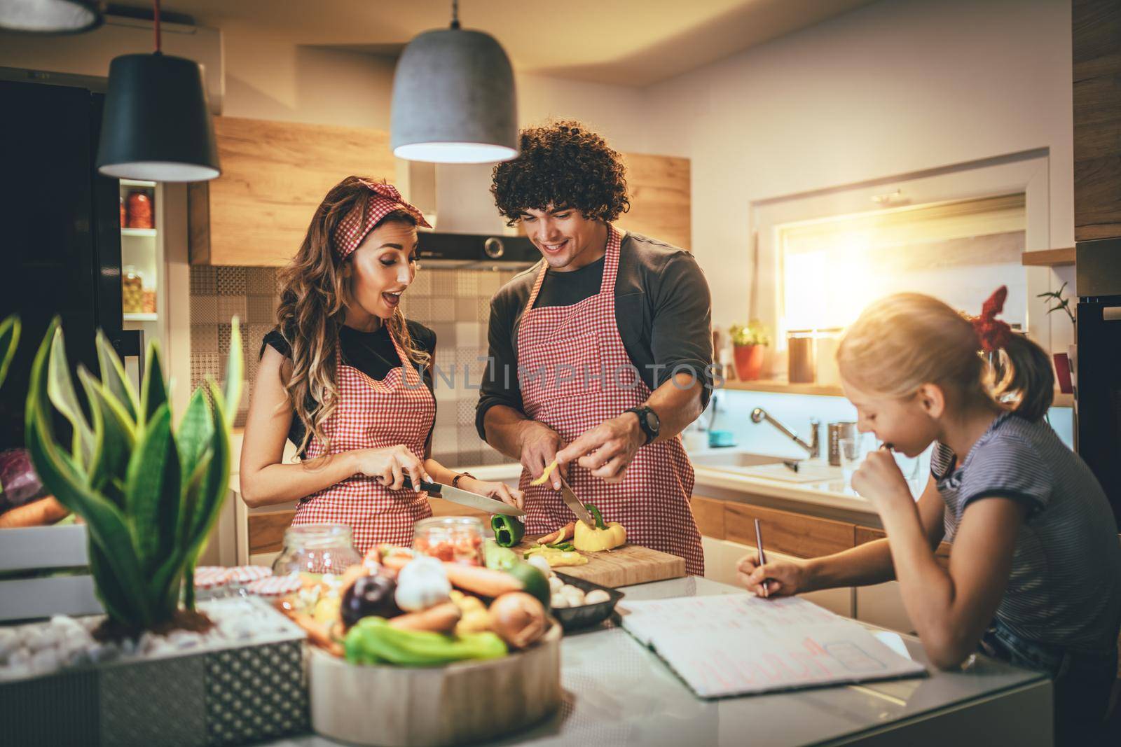 Happy parents and their daughter cooking together in the kitchen while little girl doing her homework on the kitchen table.