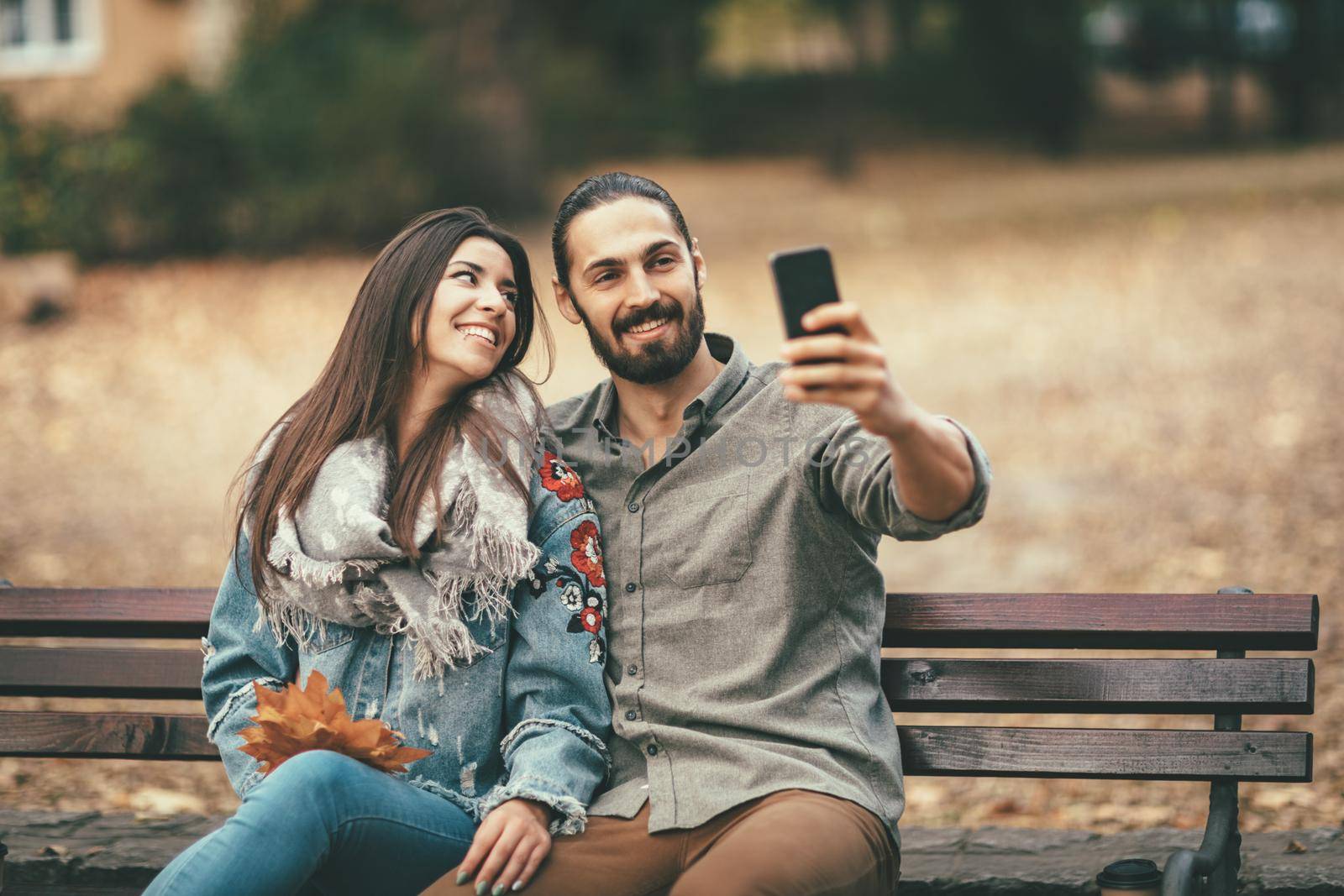 Beautiful smiling couple enjoying in sunny city park in autumn colors looking each other. They are sitting on the bench and having fun with smarthphone taking selfie.