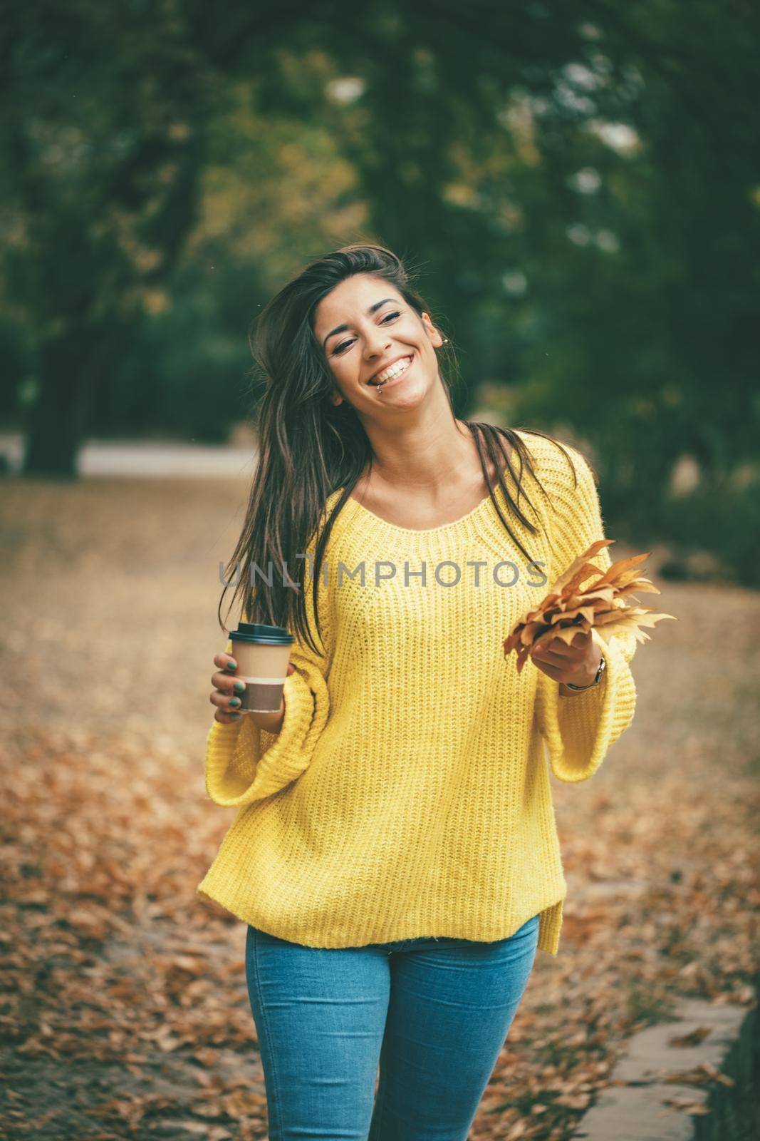 Cute young woman enjoying in sunny forest in autumn colors. She is holding golden yellow leaves and cup of cofee.