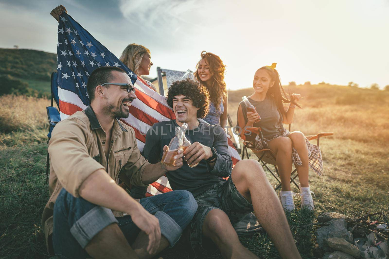 Happy young friends celebrating Independence day and enjoy a sunny day at the mountain. They're holding american flag, laughing and toasting with beer bottles in a camping.
