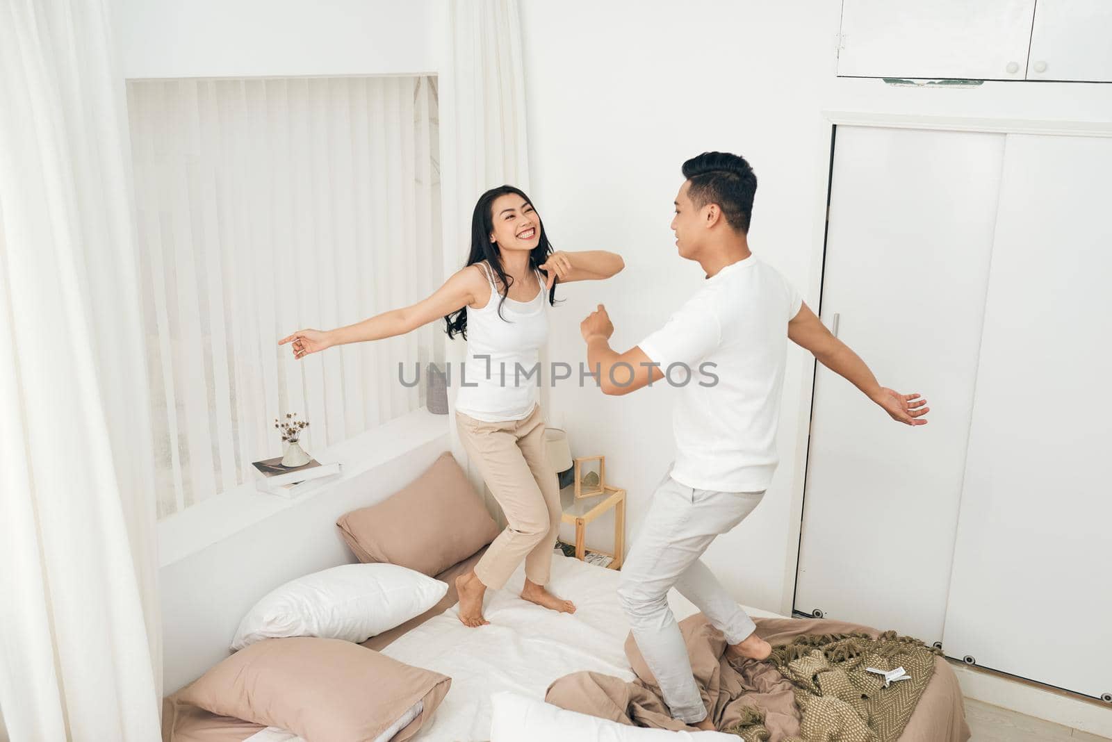 Dancing on the bed. Full length of beautiful young couple holding hands and smiling while dancing on the bed at home by makidotvn