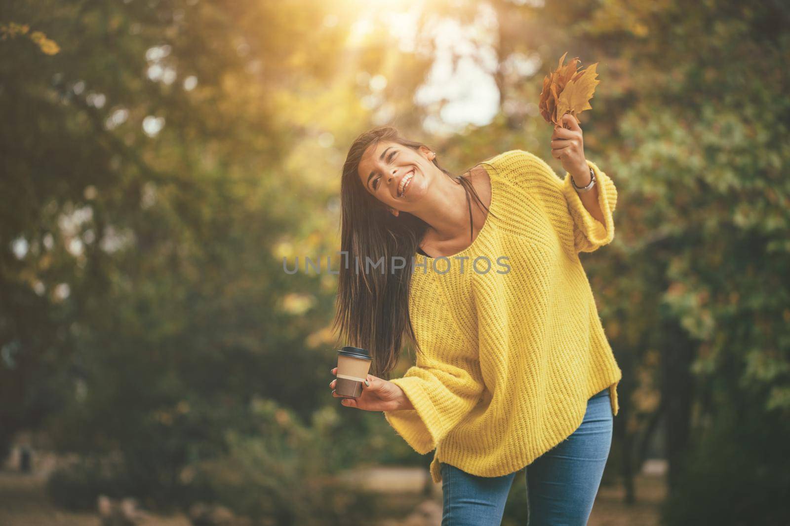 Cute young woman enjoying in sunny forest in autumn colors. She is holding golden yellow leaves and cup of cofee.