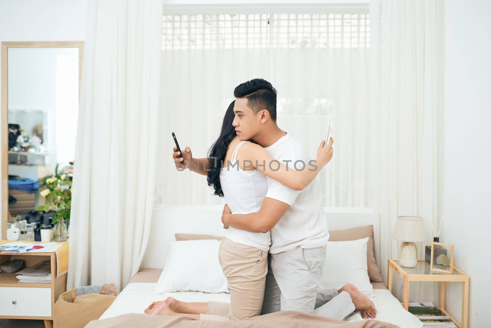 Young couple in the bedroom. Side view of unfaithful man and woman are cheating on the phone while hugging each other by makidotvn