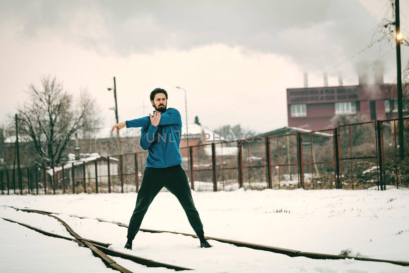 Active young man stretching and doing exercises in the old factory place among old railroad during the winter training outside in. Copy space. 