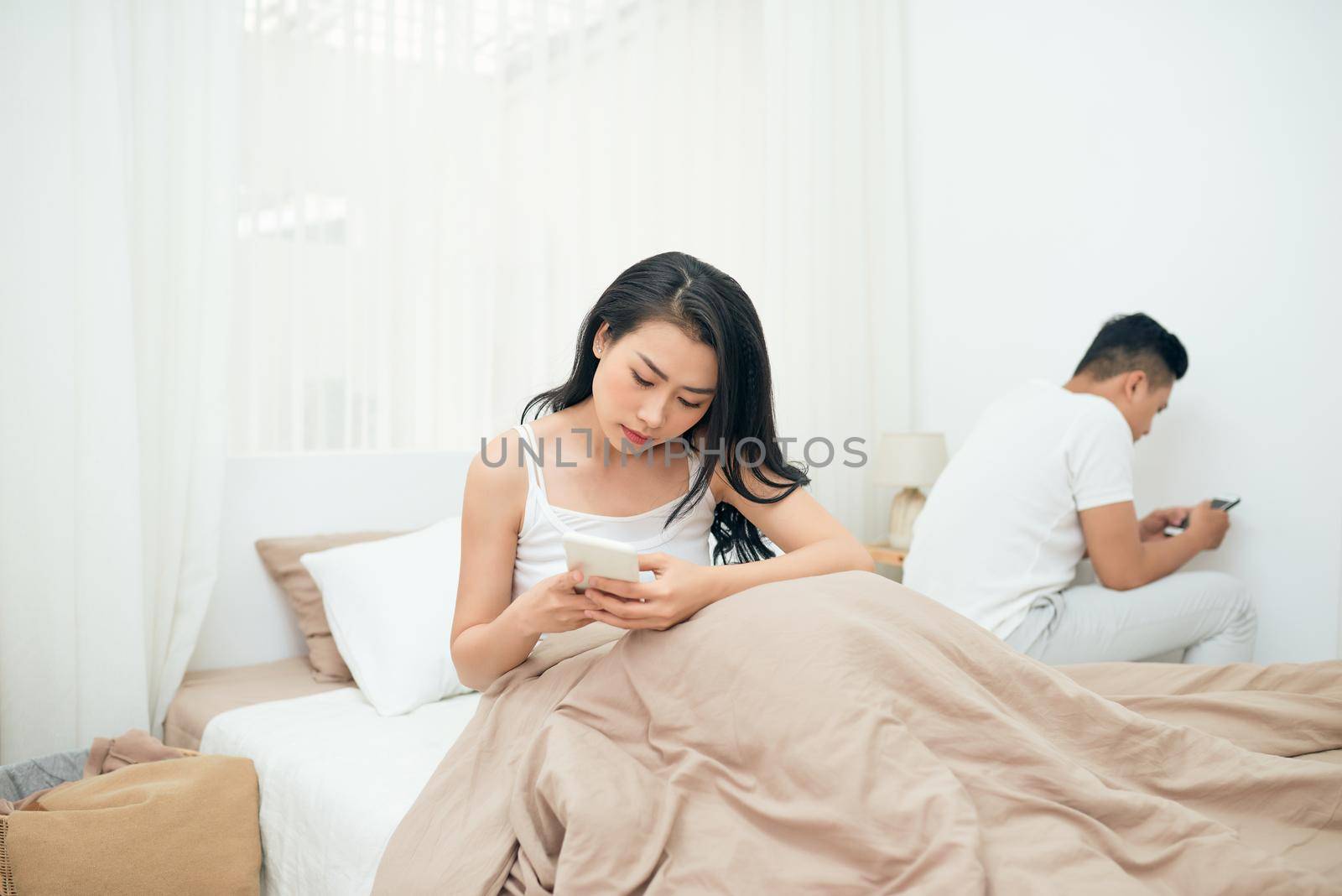 sad view of young married couple using their mobile phone in bed ignoring each other as strangers in relationship and communication problems by makidotvn