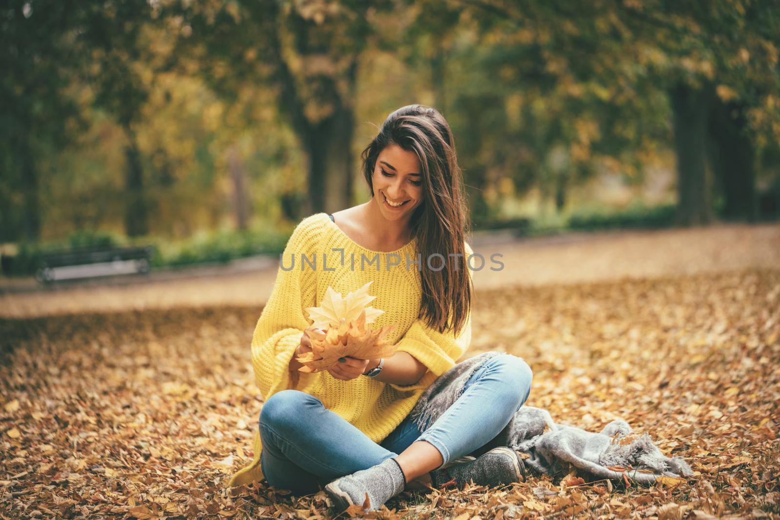 Cute young woman enjoying in sunny forest in autumn colors. She is holding golden yellow leaves and having fun. 