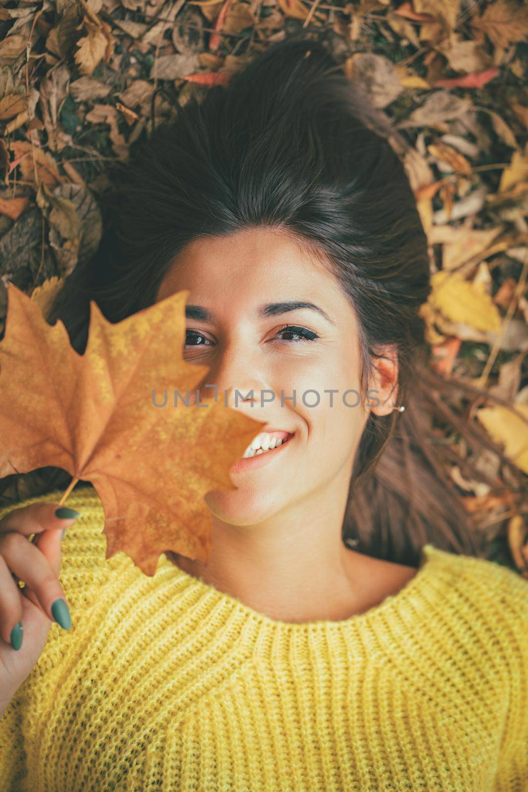 Cute young woman enjoying in sunny forest in autumn colors. She is lying down on the fall meadow looking at camera behind leaves.