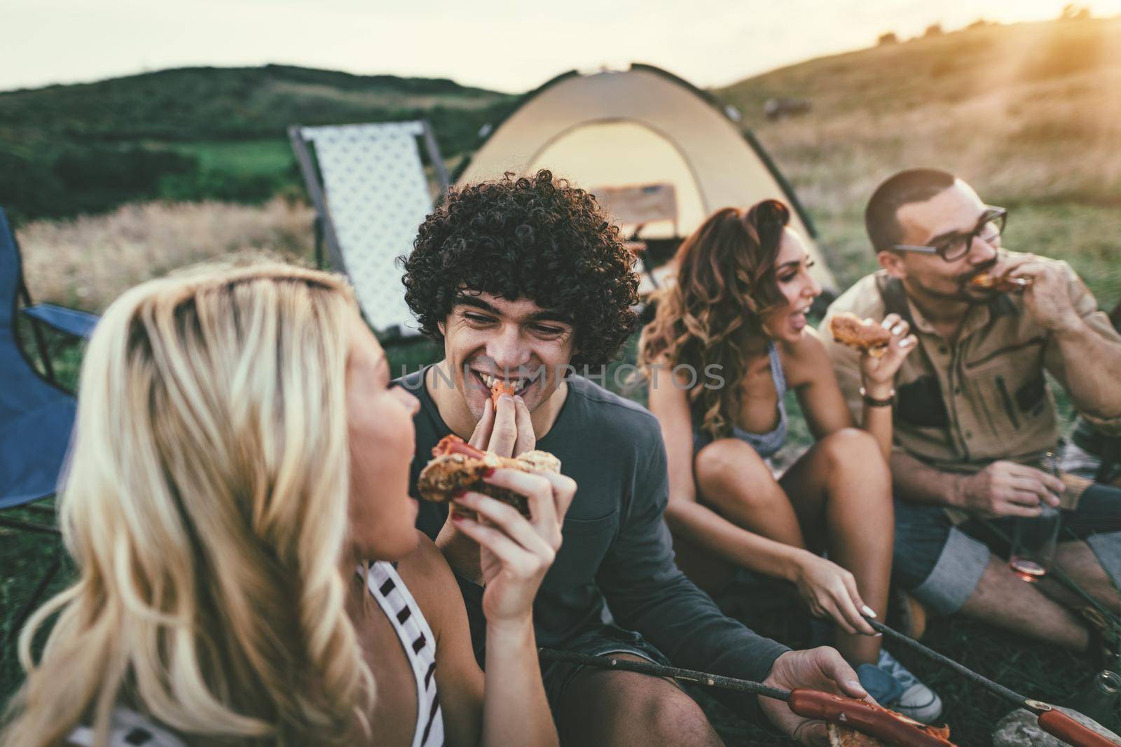 Happy young friends enjoy a nice day in nature. They're grilling sausages, eating, laughing and talking happy to be together.