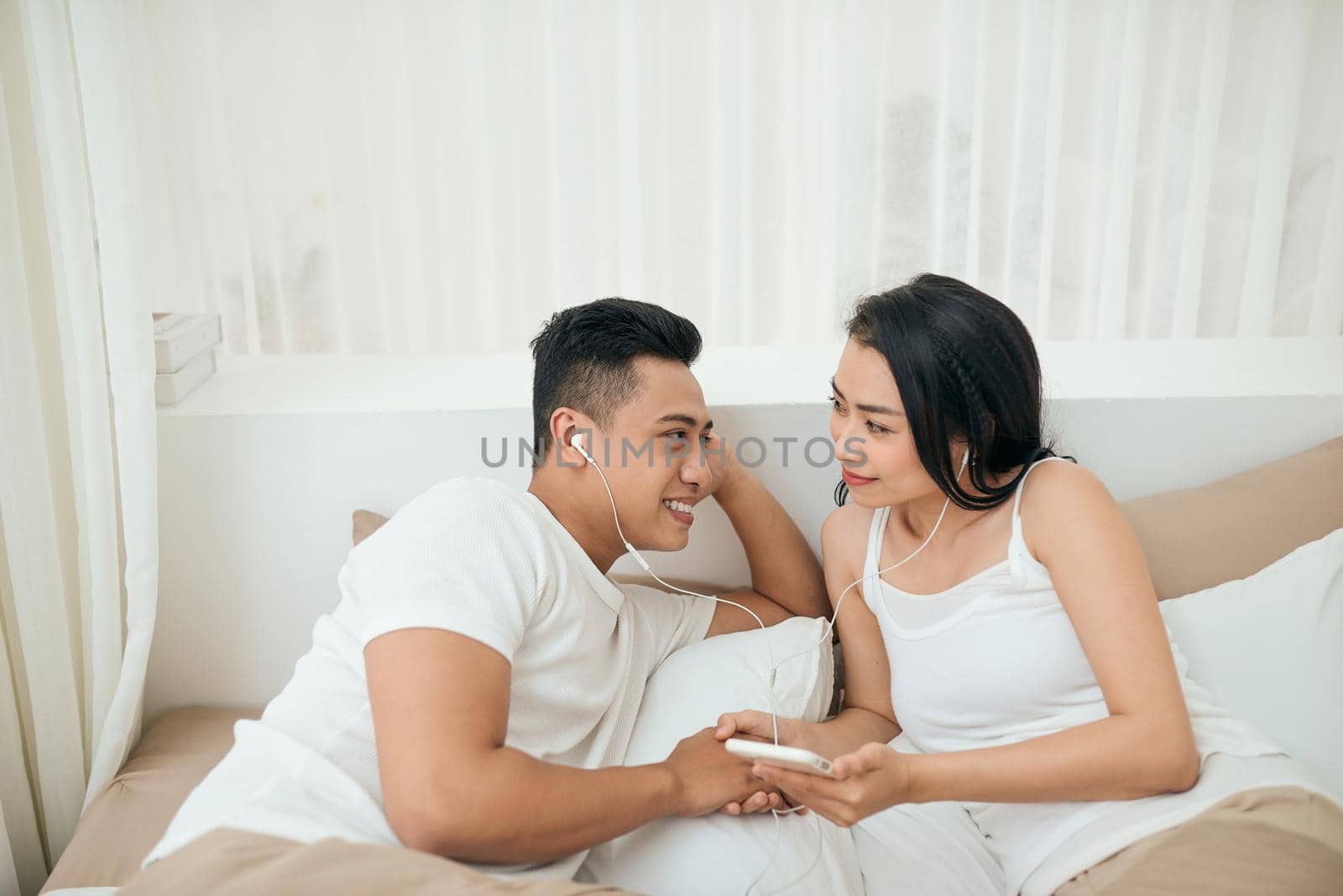 Young asian couple smile listen music with smart mobile phone on bed at bedroom with fun and enjoy