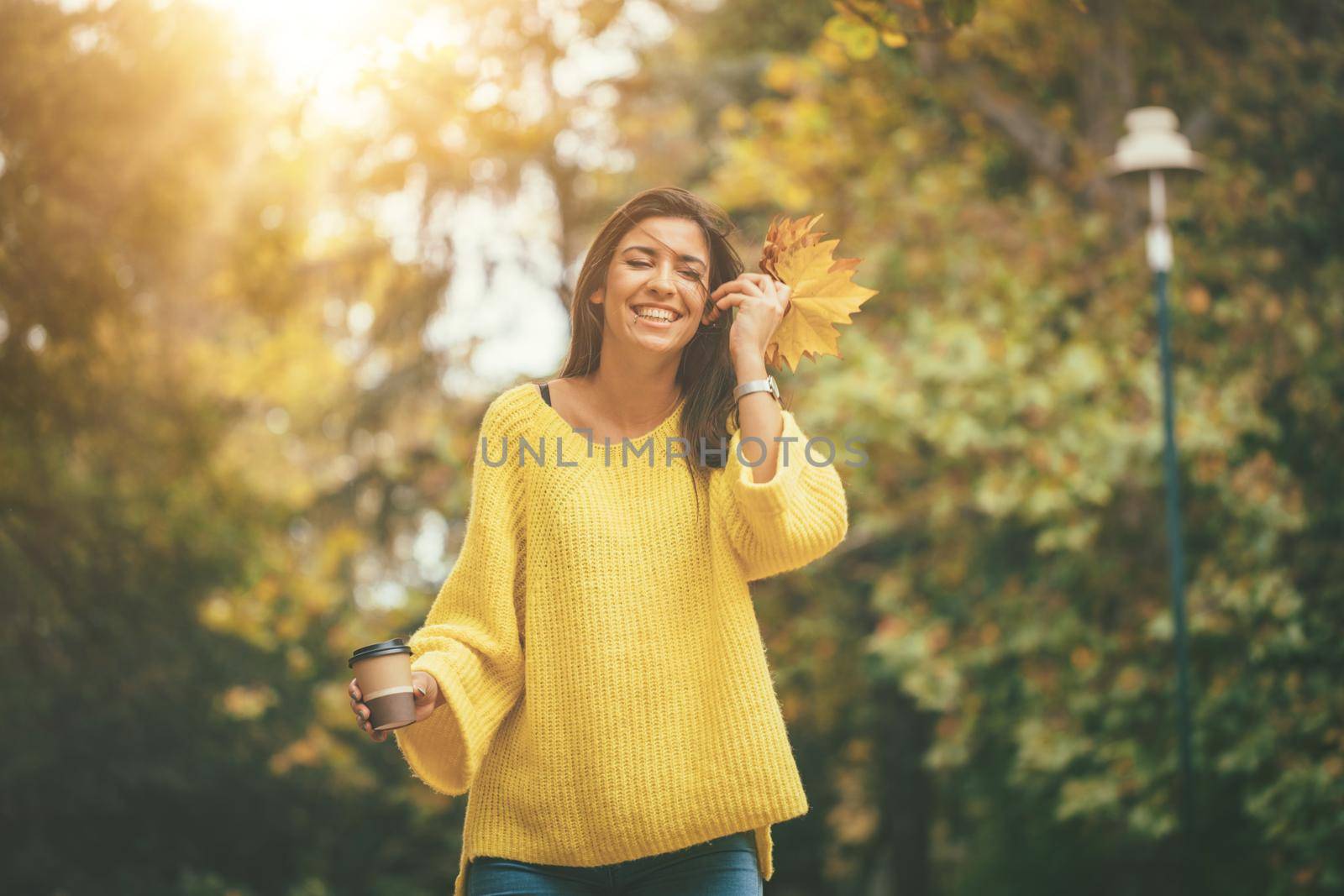 Cute young woman enjoying in sunny forest in autumn colors. She is holding golden yellow leaves and cup of cofee.