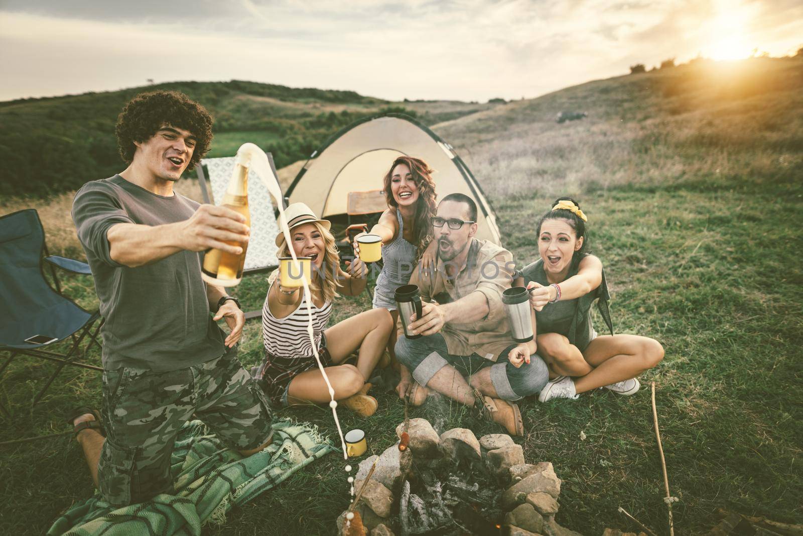 Happy young friends enjoy a nice day in nature. They're grilling sausages, eating, drinking  champagne, laughing and talking happy to be together.   