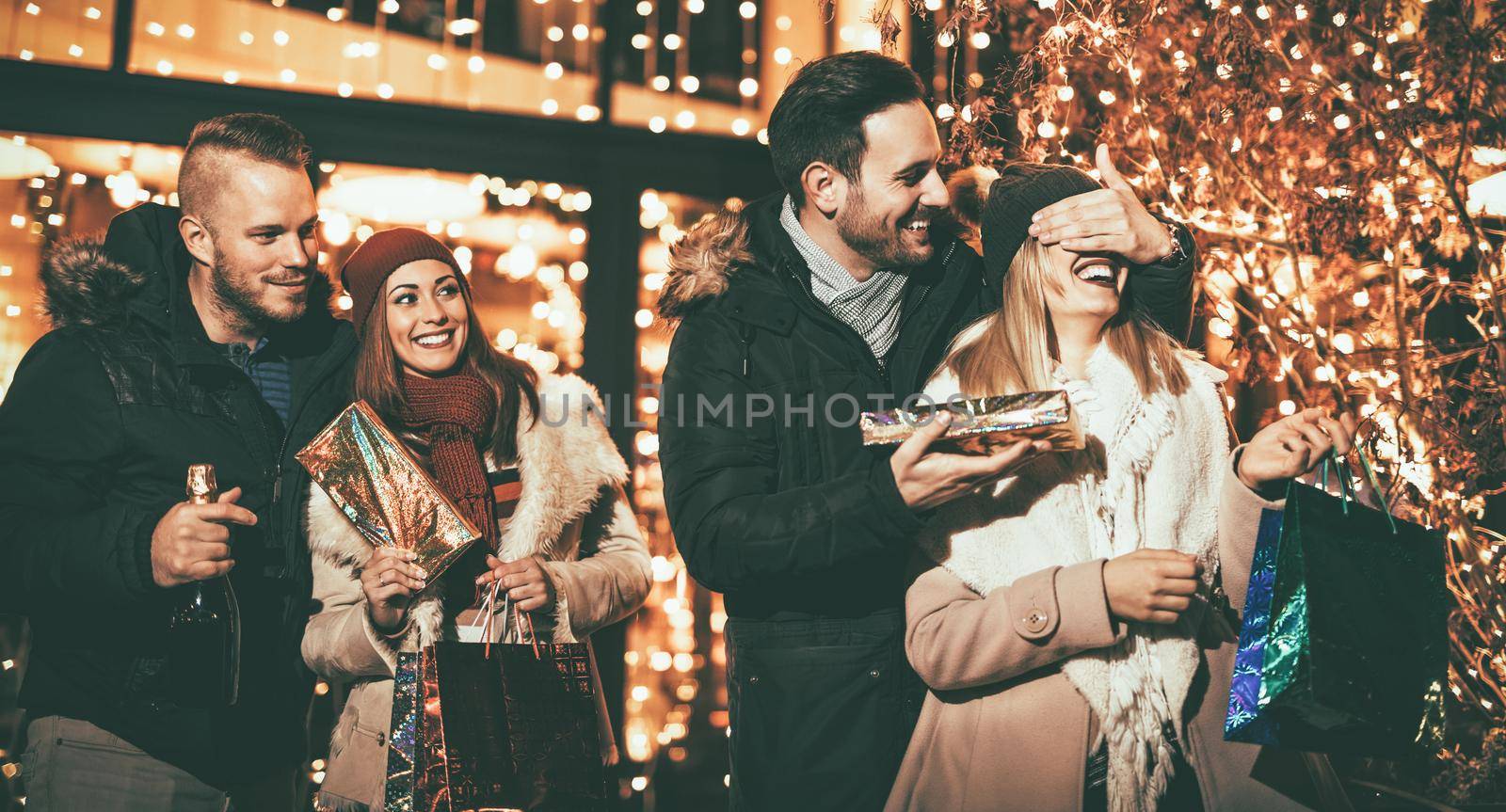 Beautiful cheerful couple celebrating Christmas in the city street. Young man covering the eyes of happy surprised girlfriend and giving her present. 