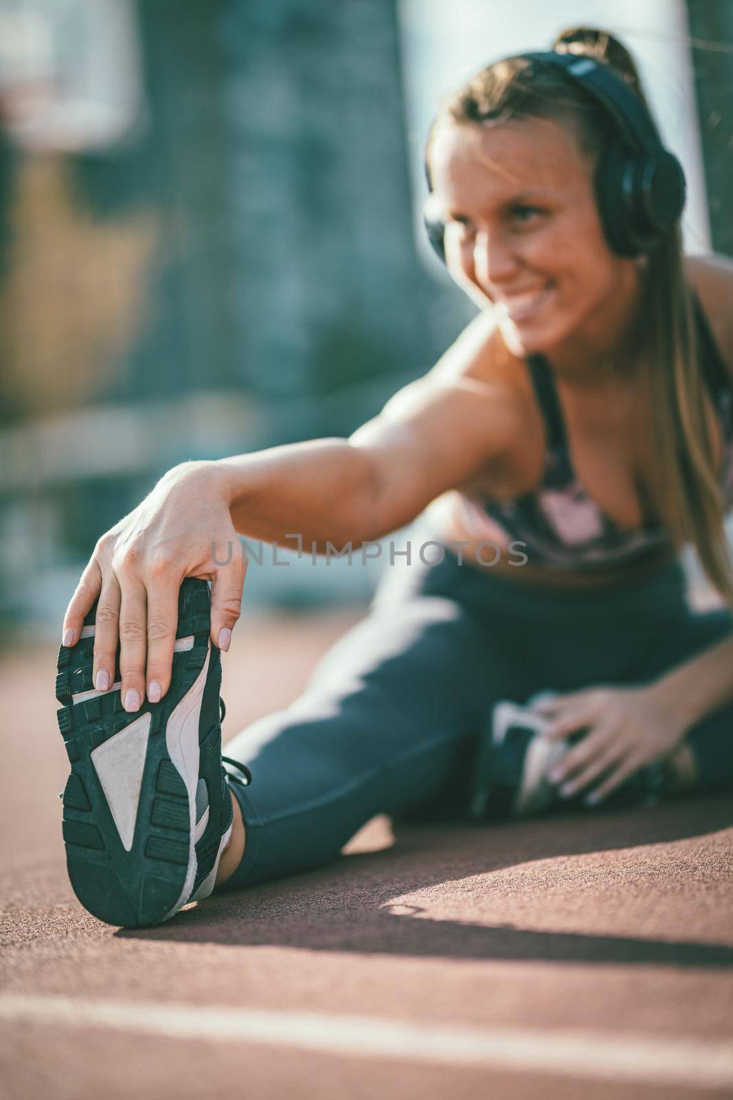 Young female runner, with earphones on her ears, doing stretching exercise on a public place, preparing for morning workout.
