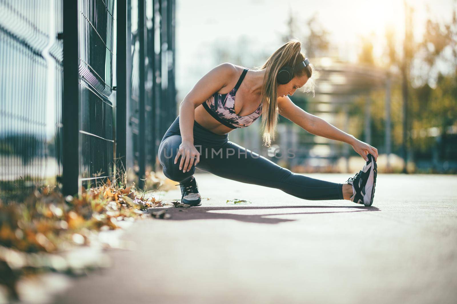 Young female runner, with headphones on her ears, doing stretching exercise on metal fence, preparing for morning workout.