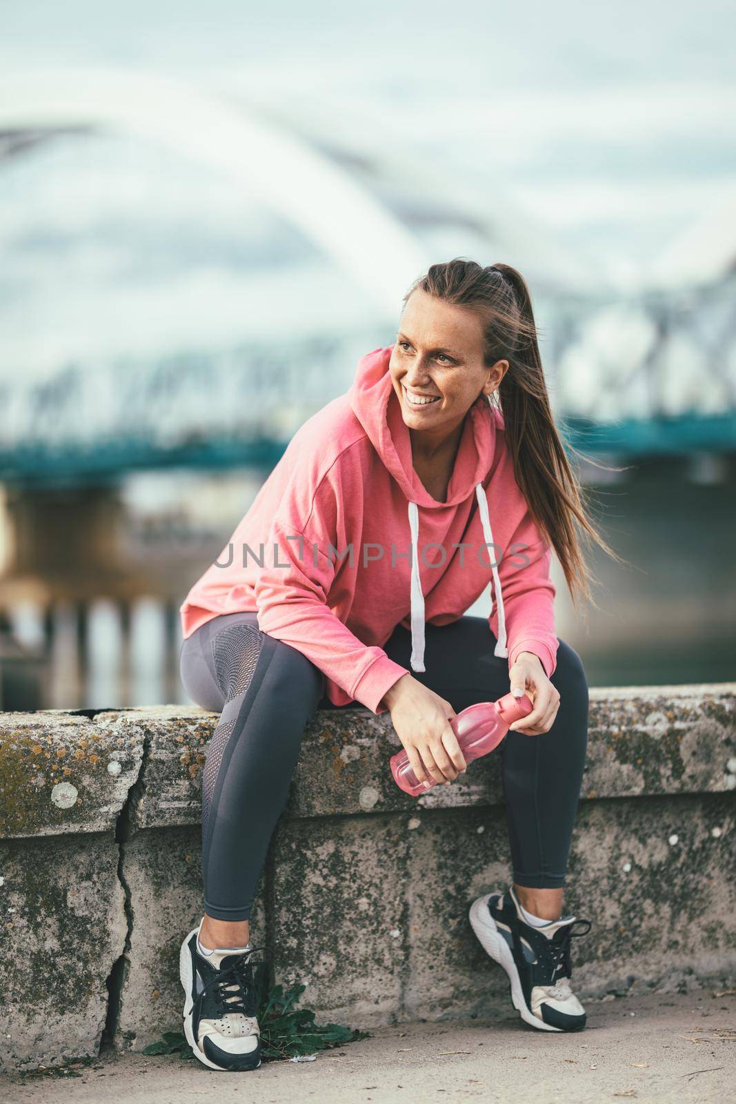 Young fitness woman is resting after hard training on the wall by the river bridge with bottle of the water.