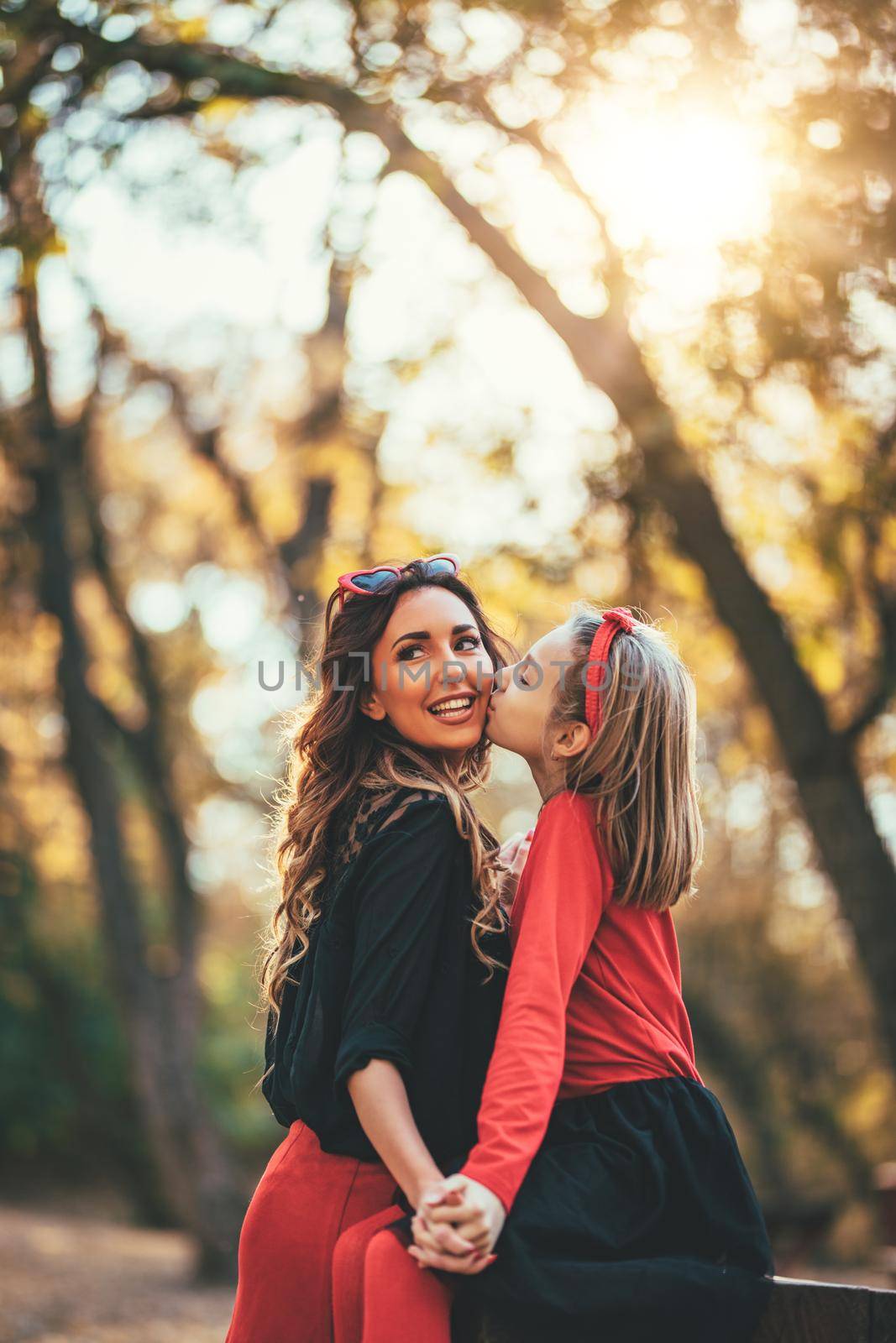 Beautiful young mother and her happy daughter having fun in the forest in sunset. Little girl is kissing mother's cheek.