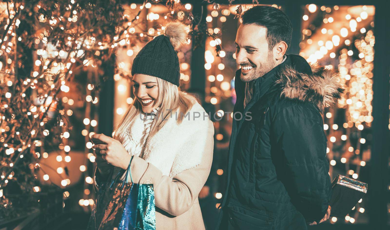 Young couple standing in shopping mall with bright shop window in background. A beautiful woman having fun while looking at smartphone.