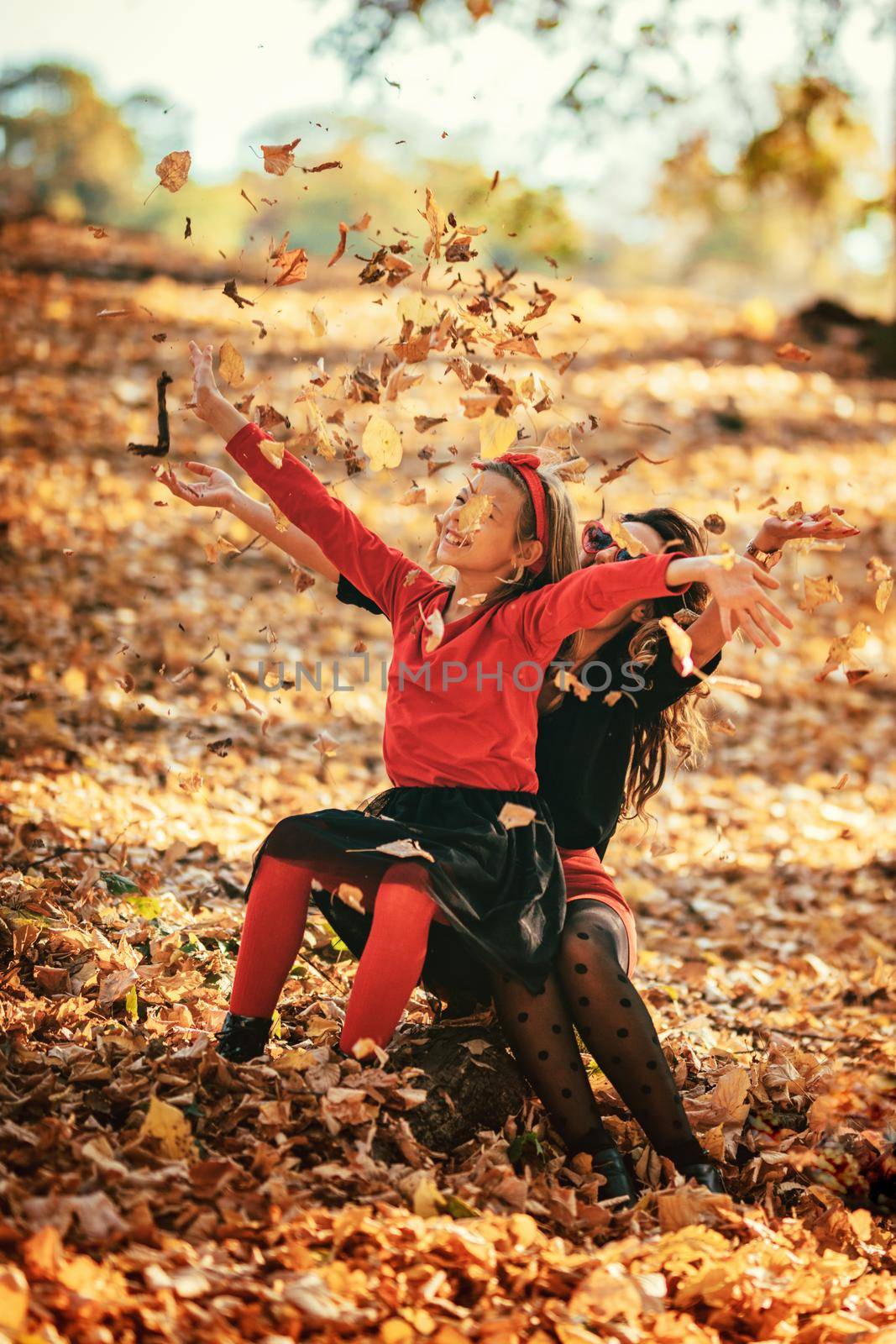 Beautiful young mother and her happy daughter having fun in the forest in sunset. They are laughing and little girl is throwing up  fallen leaves. 