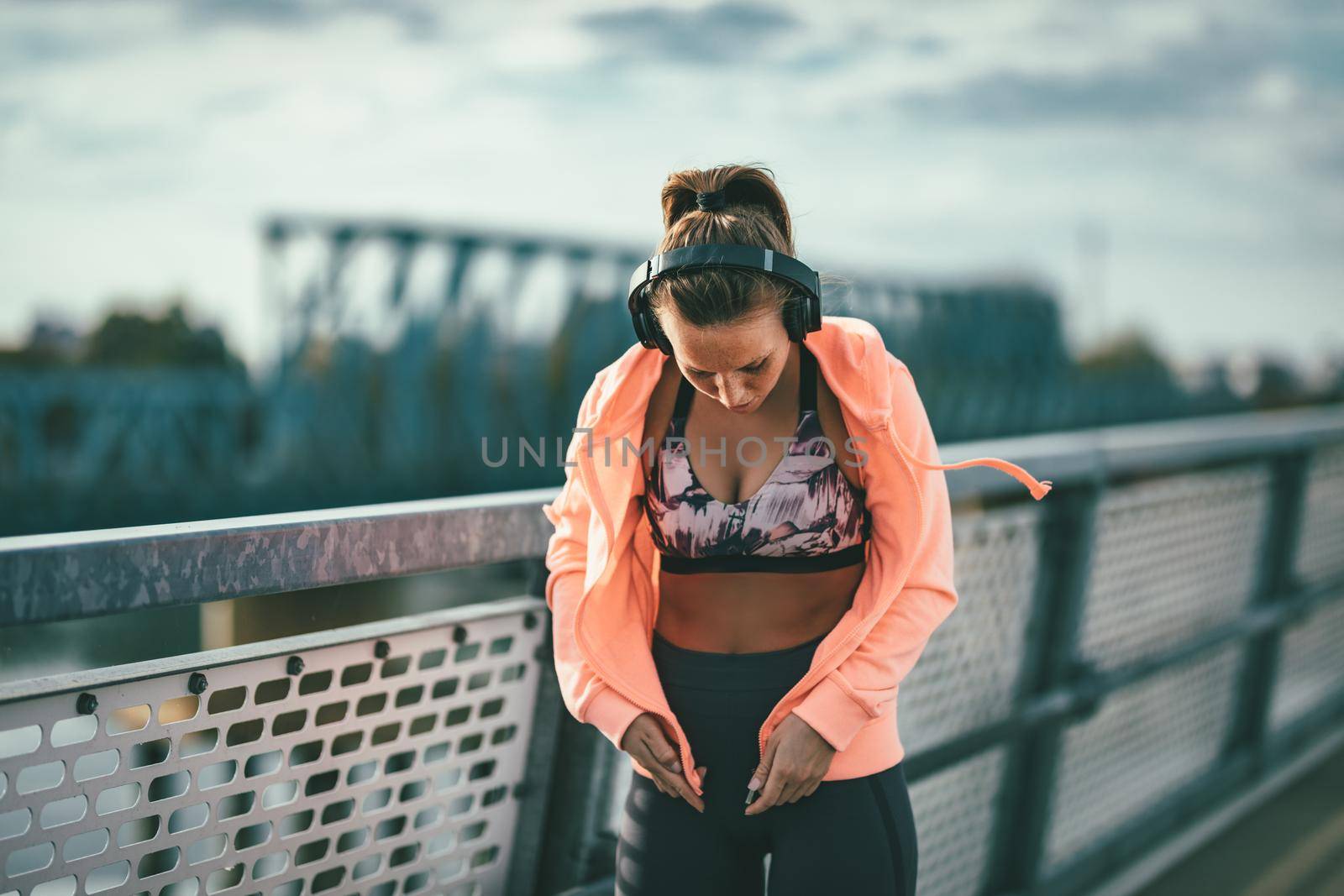 A young sport woman with headphones dresses up a sweatshirt and prepares for morning workout on a river bridge.