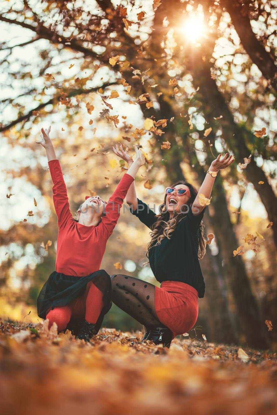 Beautiful young  mother and her happy daughter having fun in the forest in the sunset. They are crouching and having fun with fallen leaves. 