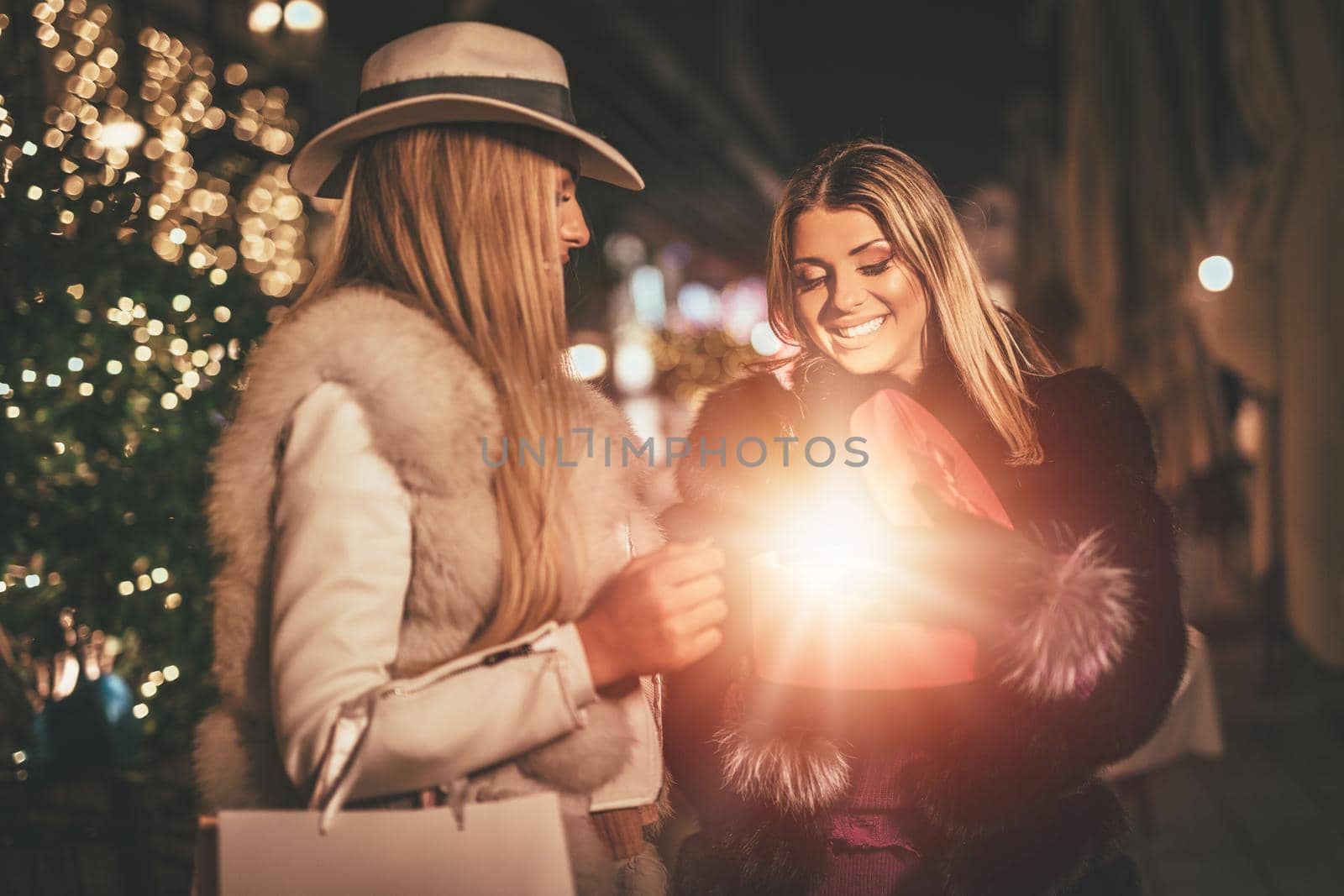 Young beautiful two cheerful sisters are celebrating Christmas on the city street, giving presents to each other. 