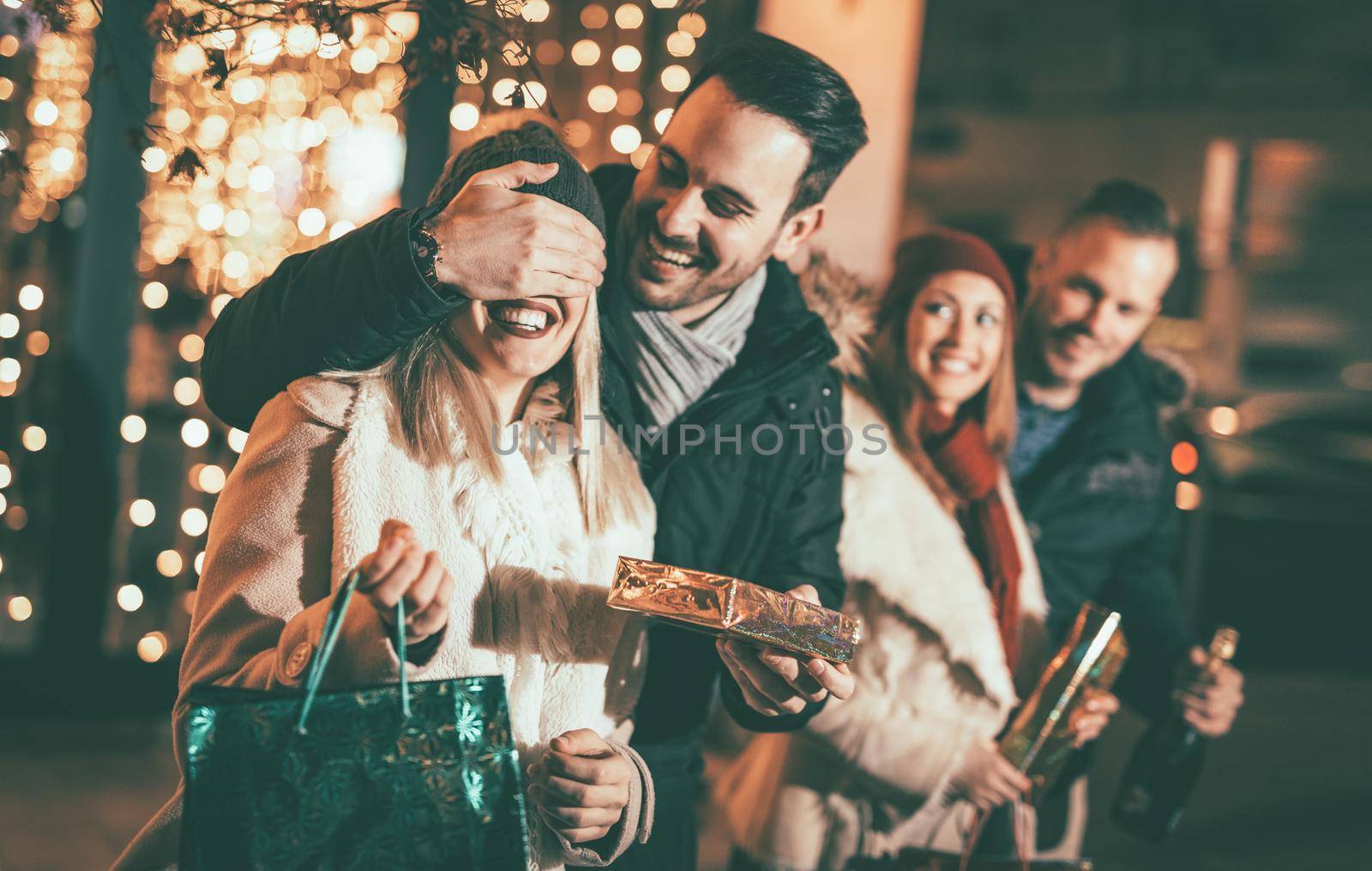 Man keeps his girlfriend eyes covered while she giving a gift. Their friends standing outside with shopping bags and boxes. Selective focus. Focus on foreground.