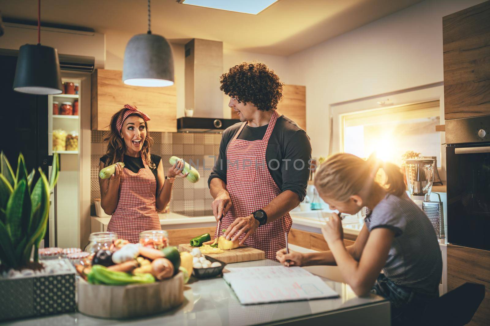 Happy parents and their daughter cooking together in the kitchen while little girl doing her homework on the kitchen table.