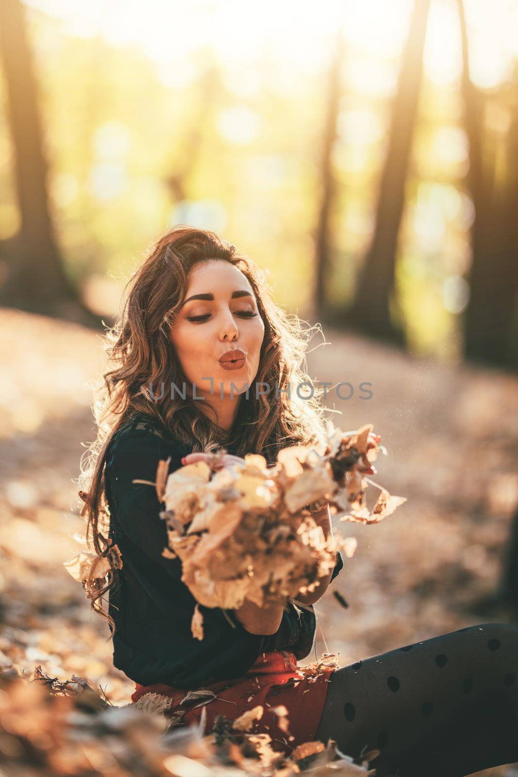 Cute young woman enjoying in sunny forest in autumn colors. She is holding golden yellow leaves, blowing on.