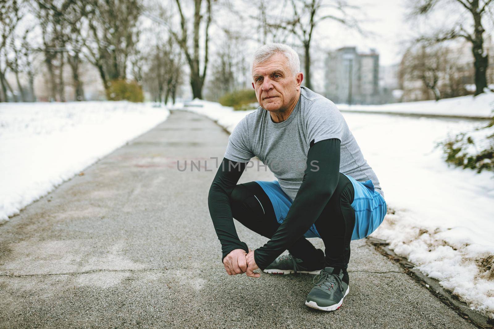 Active senior making pause crouching, during stretching and doing exercises in public park during the winter training outside in. Copy space.