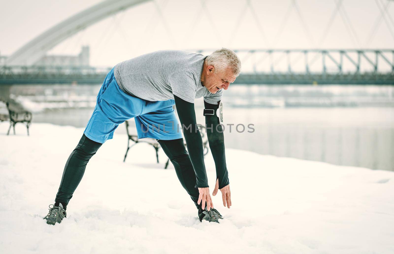 Active senior man stretching and doing exercises by the river during the winter training outside in.