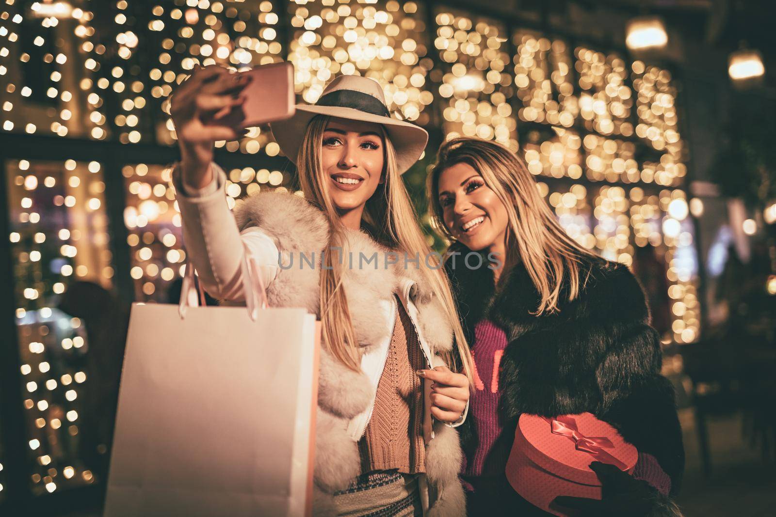 Cheerful young female friends are having fun in city street at the Christmas night. They are taking selfie with a smartphone and talking something funny.