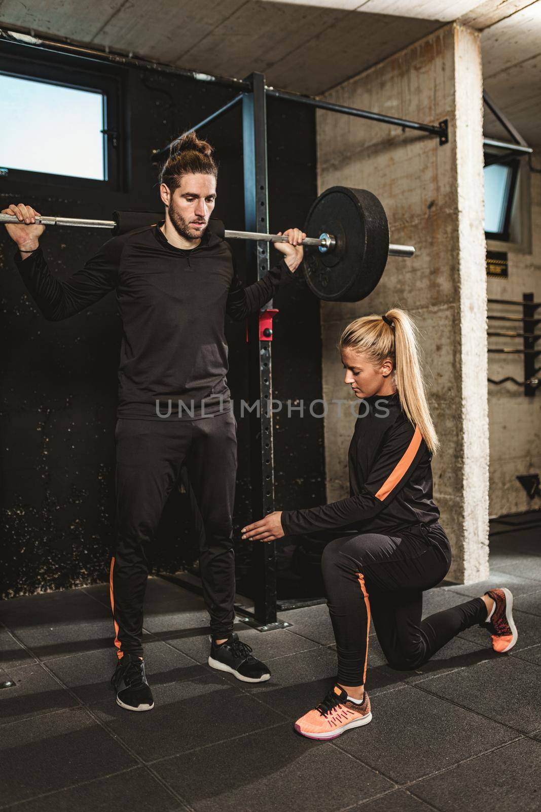 Young muscular man doing exercise with barbell at the gym, with support of his personal female trainer. 