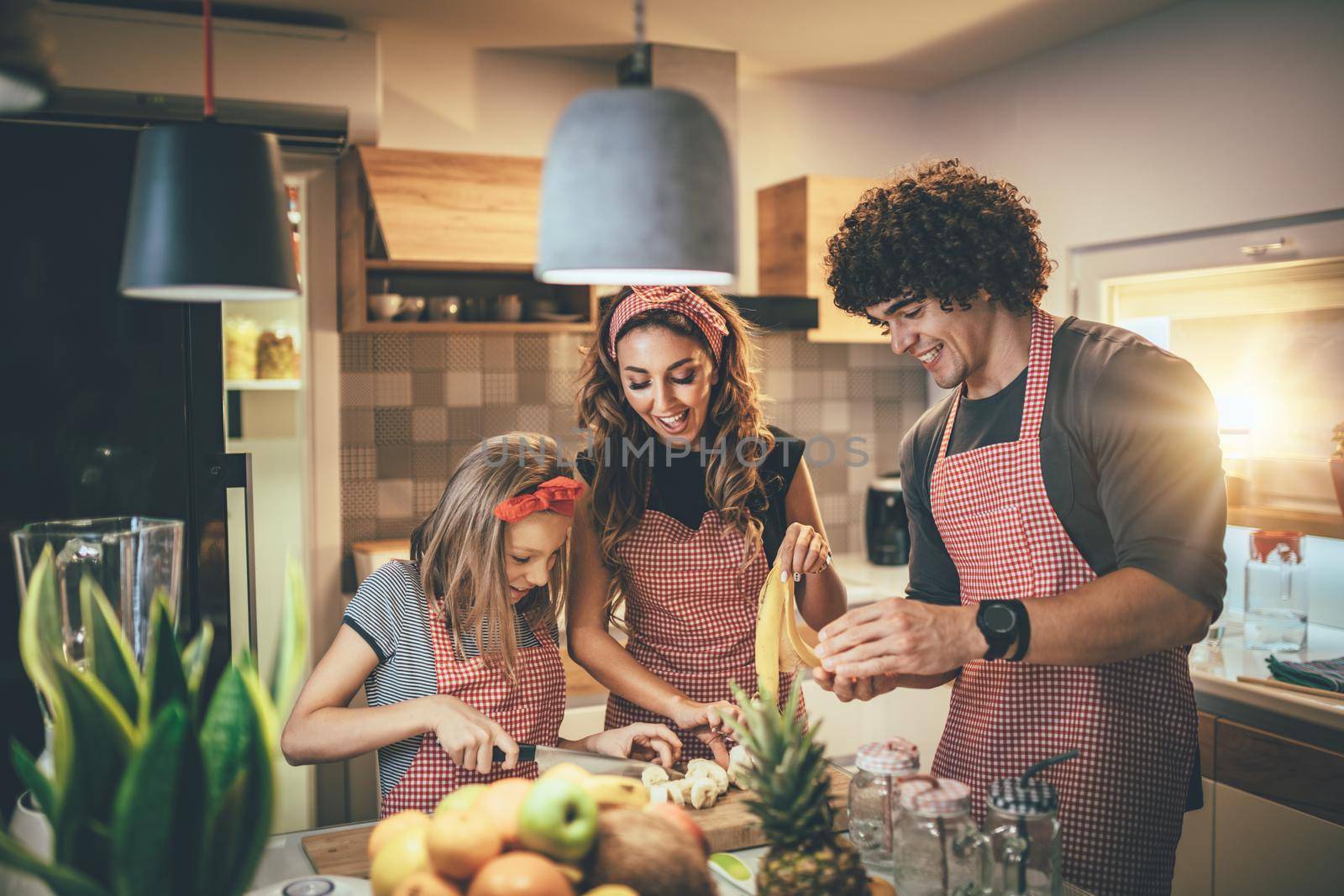 Happy parents and their daughter preparing healthy meal together in the kitchen while little girl trying to cut banana.