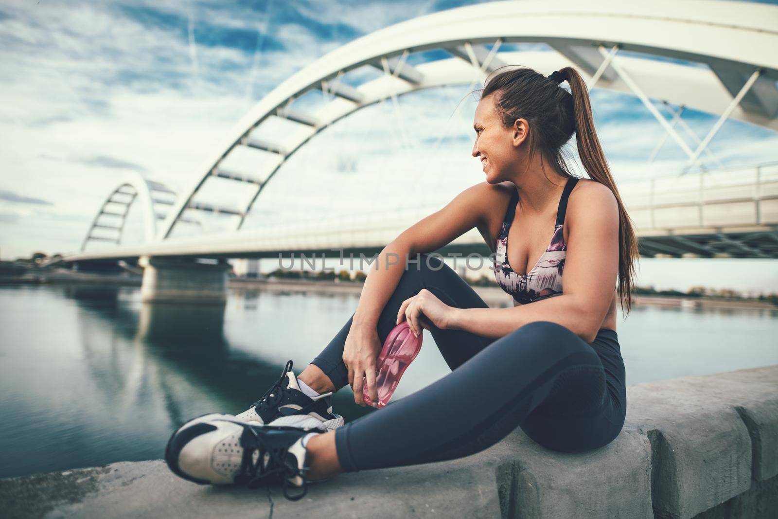 Young fitness woman is resting after hard training by the river bridge drinking water.