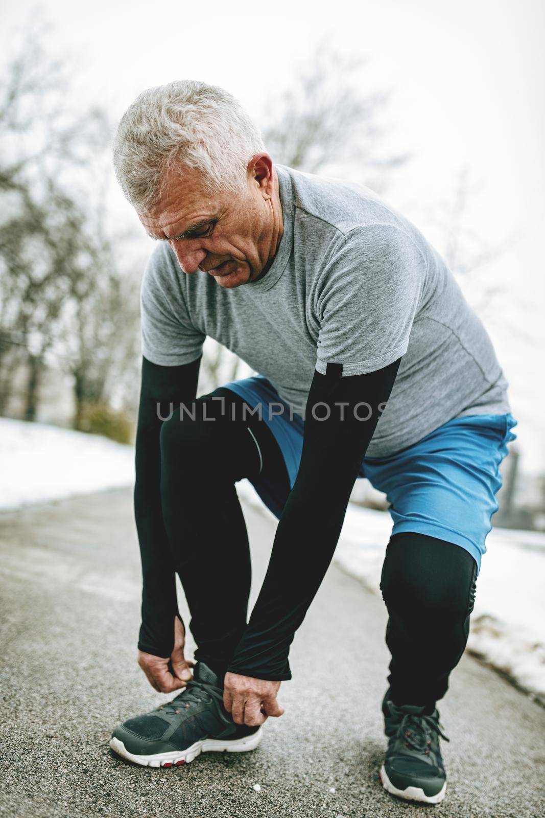 Active senior tying running shoes, and making pause during jogging and doing exercises in the public park in the winter training outside in.