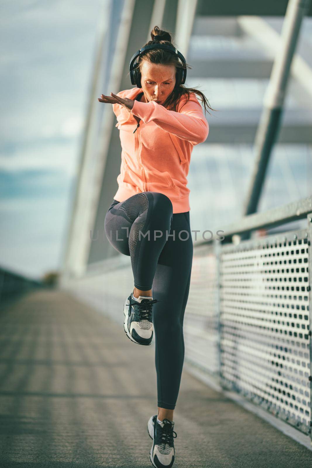 Young fitness woman with headphones doing warm up exercise on river bridge, preparing for morning workout.