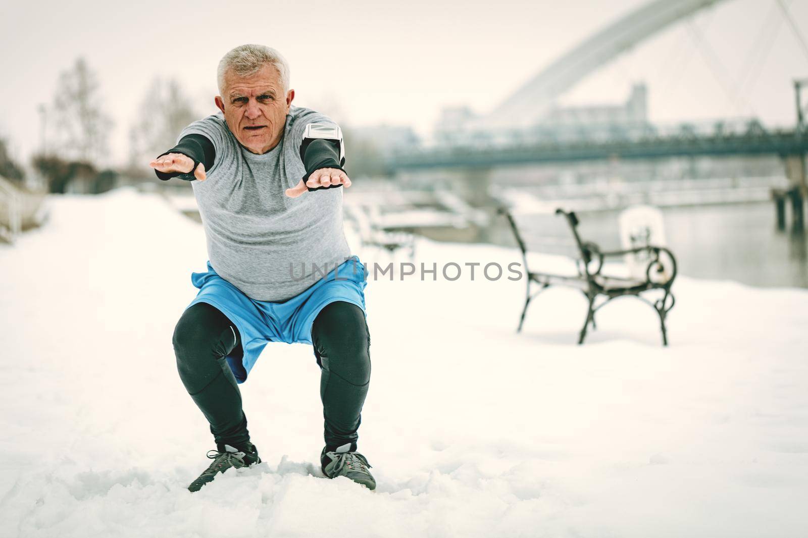 Active senior man stretching and doing squats by the river during the winter training outside in. Copy space.