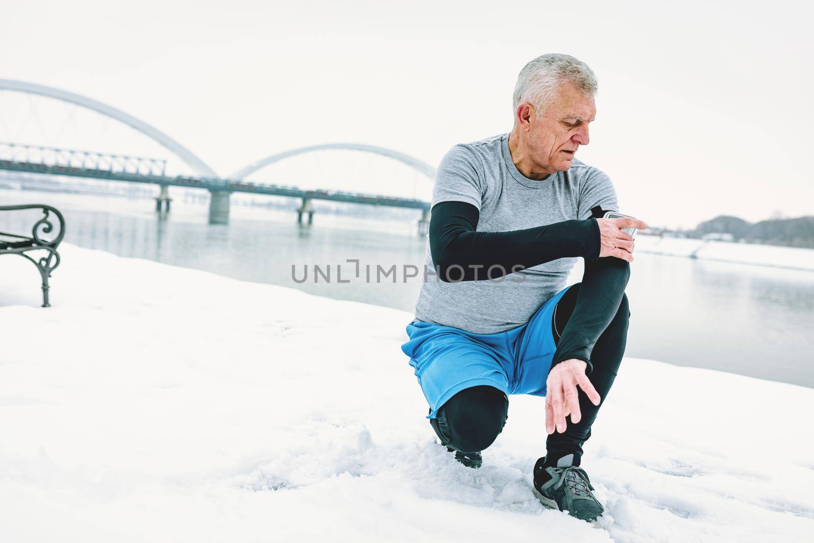 Active senior man checking mobile phone during doing exercises by the river in the winter training outside in. Copy space.