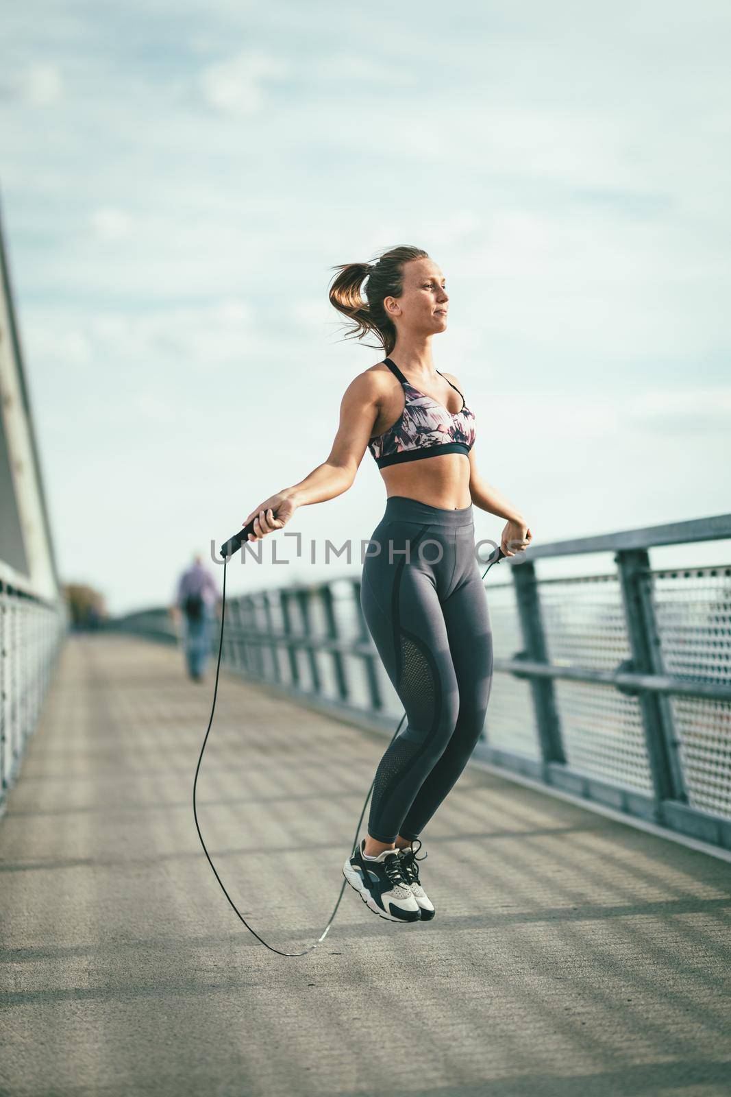 Young female runner jumping over skipping rope and doing exercise on a river bridge, preparing for morning workout.