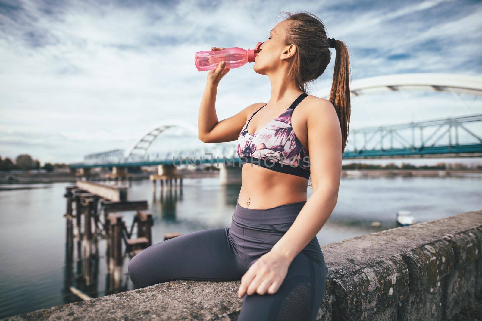 Young fitness woman is resting after hard training on the wall by the river bridge and drinking water.