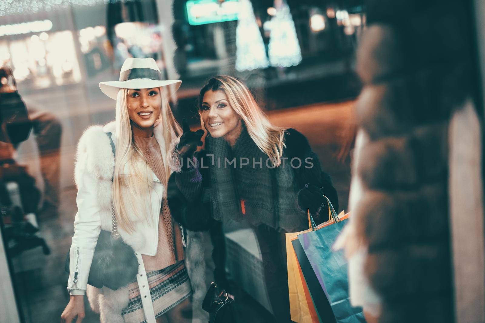 Young beautiful cheerful two sisters having fun in the city street at New Year time and pointing at something through the window shop.