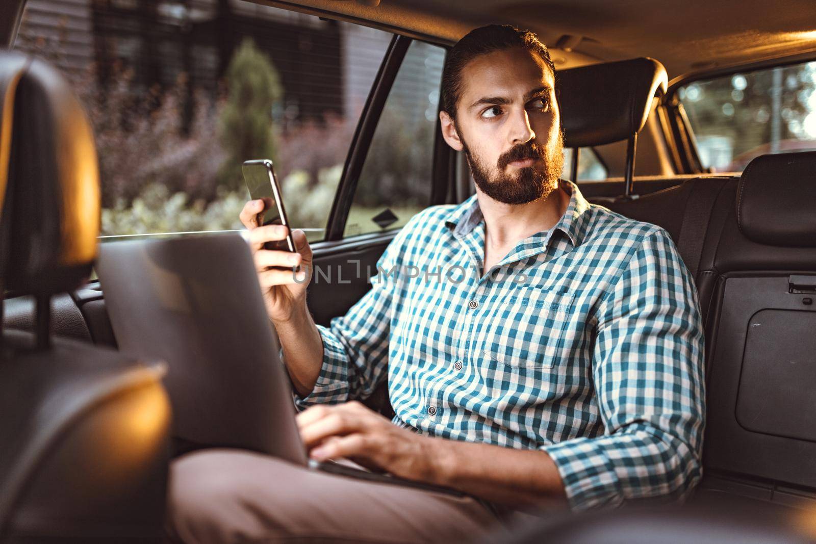 Young confident businessman is holding smartphone, talking and working on a laptop on the back seat in car.