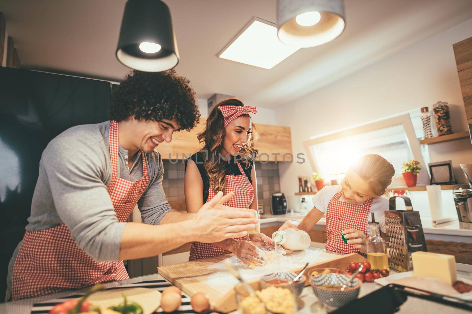 Happy parents and their daughter are preparing dough for pizza together in the kitchen. Little girl helps to her parents to pour milk in the bunch of flour on the table.