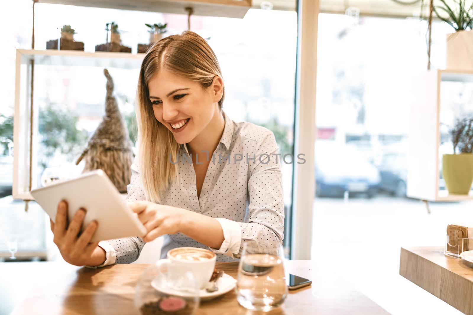 Beautiful young smiling woman drinking coffee in a cafe. She is surfing the internet on a digital tablet.