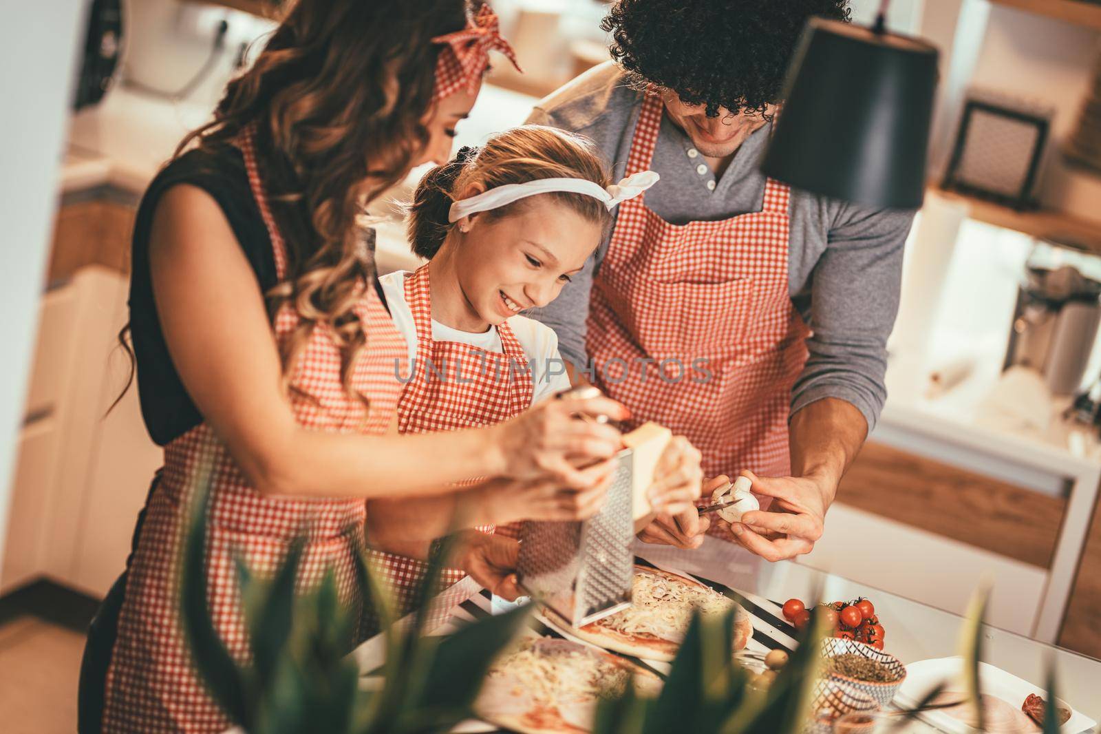 Happy parents and their daughter are preparing meal together in the kitchen. Little girl and her mother are putting cheese on the pizza and her father is cutting mushrooms.