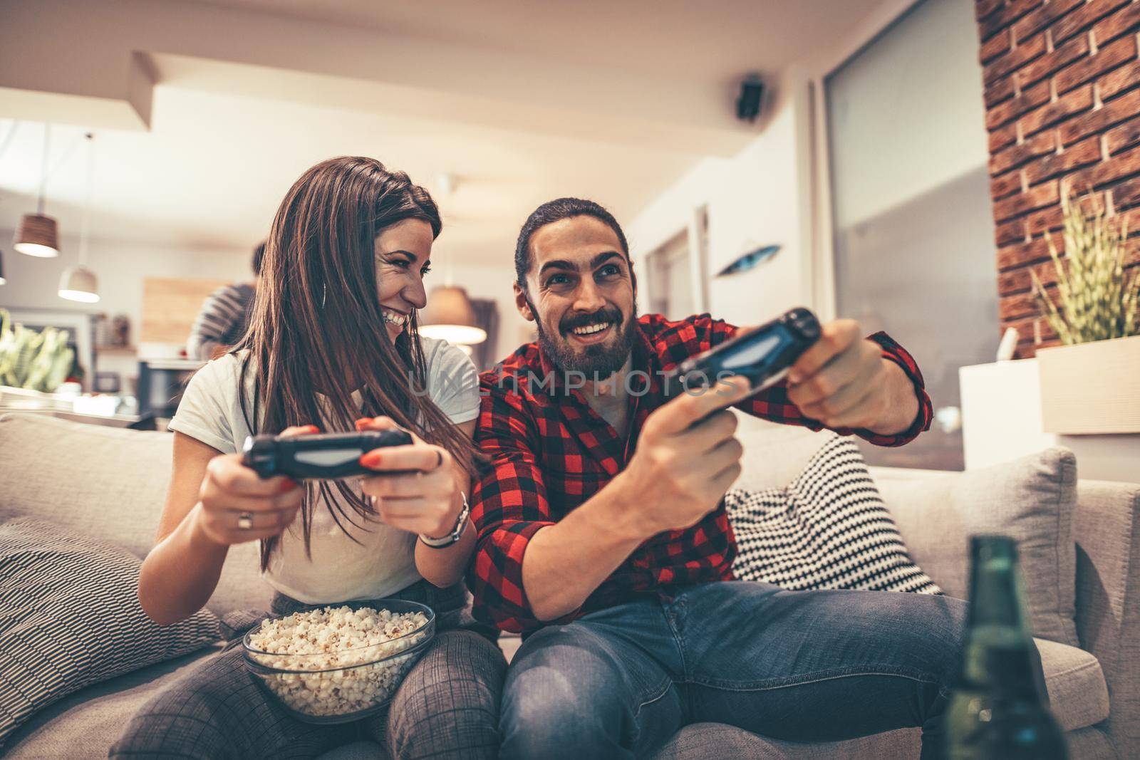 Excited couple is playing video games at home sitting on sofa with popcorn. They have great weekend in nice company indoors. 